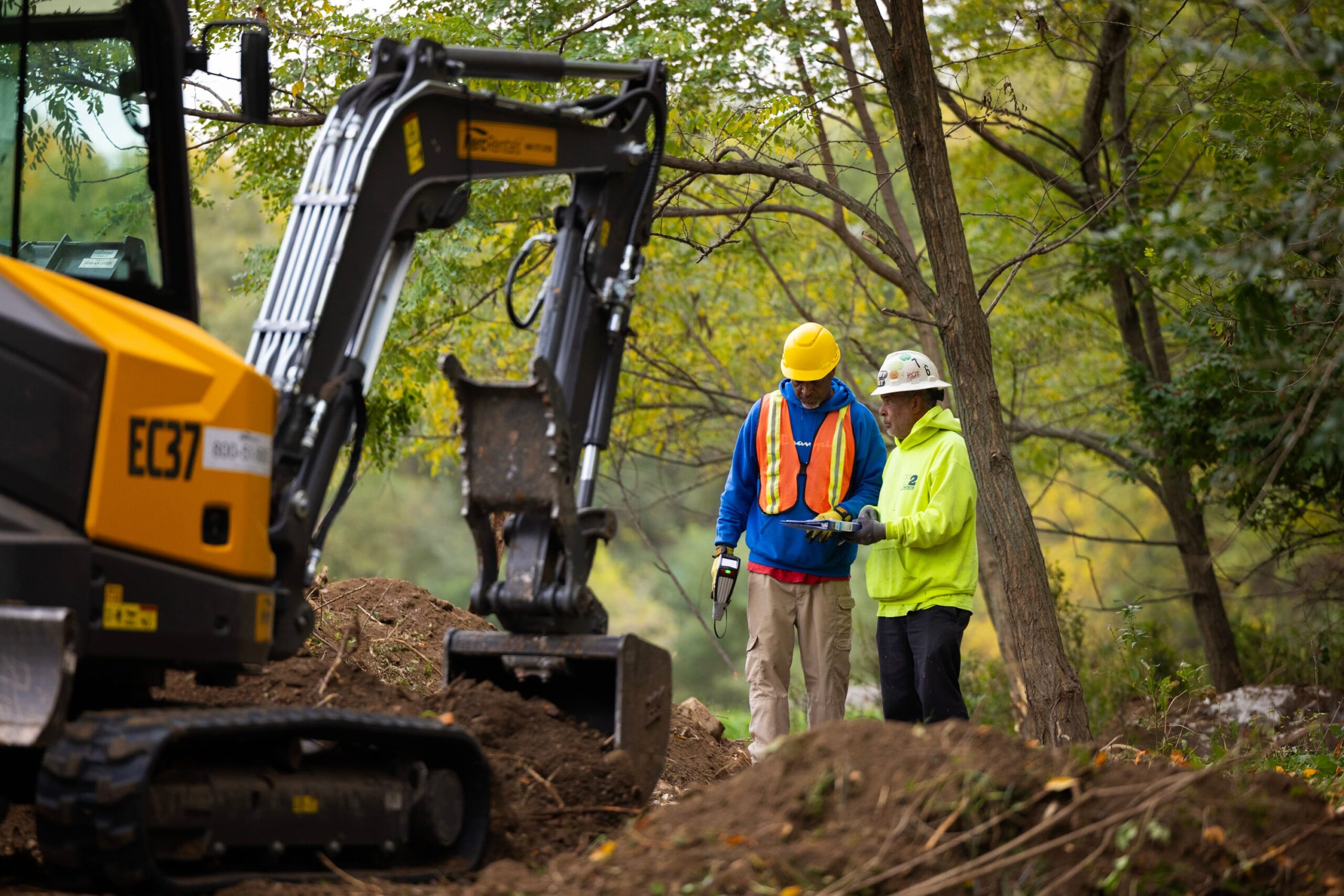 Two men in hard hats and fluorescent jackets stand next to a backhoe