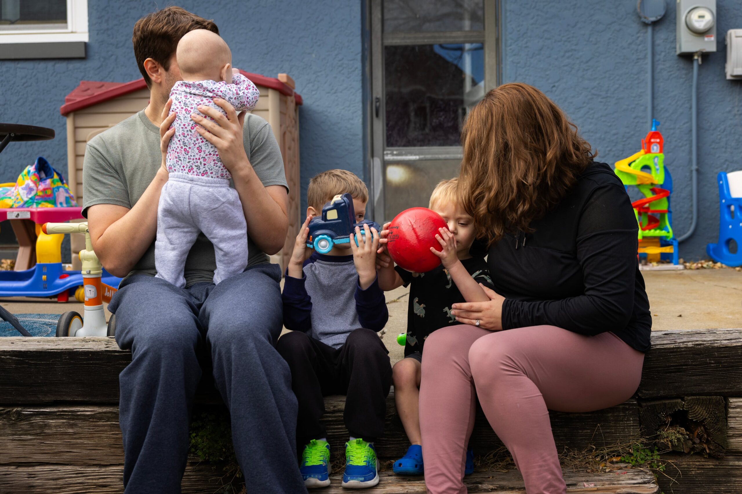A woman and man play with a baby and toddler in front of a house